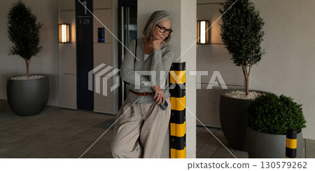Woman in stylish attire leaning against a column in a modern building during the day with plants 130579262