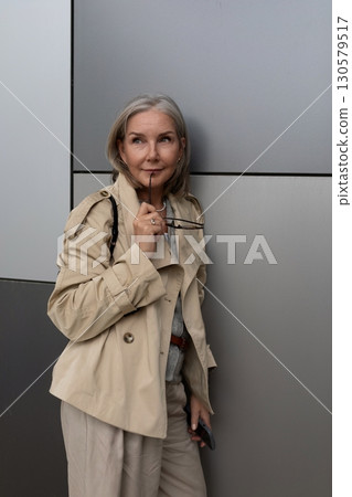 Mature woman in stylish trench coat poses thoughtfully against modern architecture in a vibrant Mature woman in stylish trench coat poses thoughtfully against modern architecture in a vibrant 130579517
