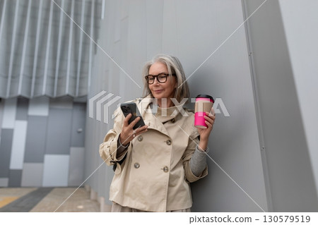 Woman in stylish trench coat checks her smartphone while enjoying a beverage outside a modern Woman in stylish trench coat checks her smartphone while enjoying a beverage outside a modern 130579519
