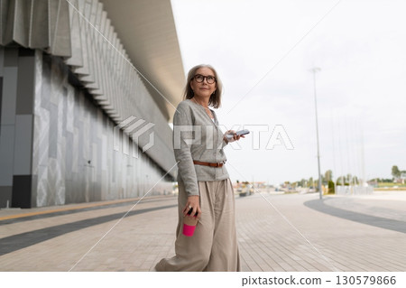 Mature woman walking with coffee cup and smartphone near modern architecture in urban area Mature woman walking with coffee cup and smartphone near modern architecture in urban area 130579866