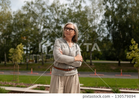 Senior woman stands confidently in a park while enjoying a sunny day outdoors surrounded by green Senior woman stands confidently in a park while enjoying a sunny day outdoors surrounded by green 130579894