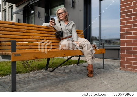 Elderly woman relaxing on a bench using her smartphone in an urban outdoor setting during daytime Elderly woman relaxing on a bench using her smartphone in an urban outdoor setting during daytime 130579960