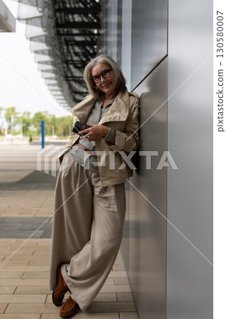 Elegant older woman relaxed and smiling while using smartphone near modern building in bright Elegant older woman relaxed and smiling while using smartphone near modern building in bright 130580007