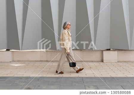 Senior woman walks confidently along modern architecture in urban setting under bright daylight 130580086