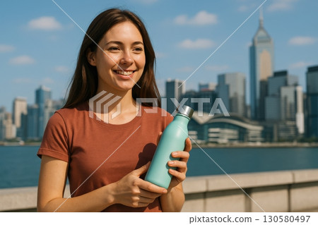 Young woman enjoying a refreshing drink with a city skyline backdrop in bright daylight 130580497