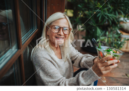 Senior woman standing terrace, eating healthy dinner outdoors. 130581314