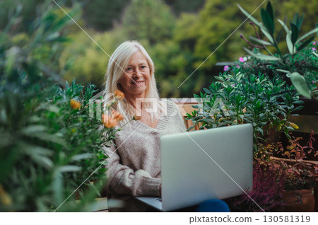 Mature woman sitting on balcony with laptop, working remotely. 130581319
