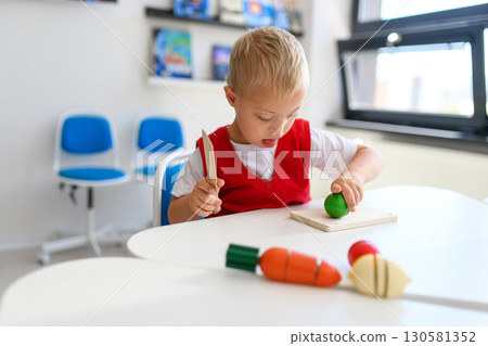 Boy with down syndrome cutting toy vegetables in classroom. Boy with down syndrome cutting toy vegetables in classroom. 130581352