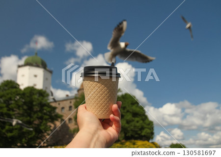 Brown paper cup of drink in women's hand on against backdrop of island Olaf's Tower in town Vyborg and flying seagull in background in blur, Russia on sunny day 130581697