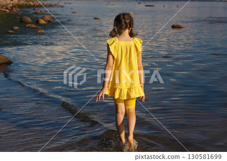Little girl walking along seashore in yellow dress on sunny day, view from back 130581699