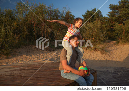Happy little cheerful daughter on daddy's back with arms to sides playing happy in airplane on sunny beach in summer, lifestyle, , Father's Day 130581706