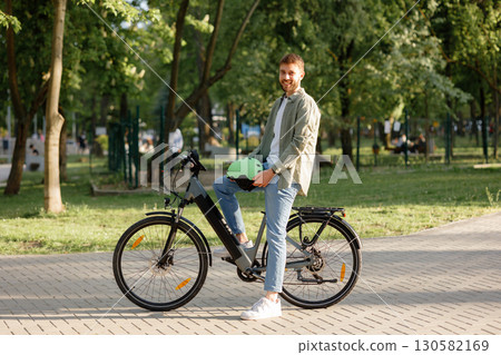 Smiling man enjoying a sunny day while posing with his electric bike in a park setting Smiling man enjoying a sunny day while posing with his electric bike in a park setting 130582169