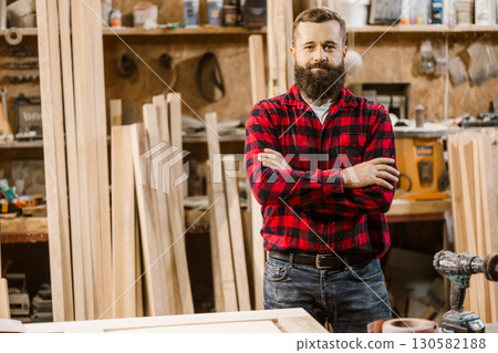 Skilled carpenter poses confidently in a woodworking workshop surrounded by tools and wooden materials Skilled carpenter poses confidently in a woodworking workshop surrounded by tools and wooden materials 130582188