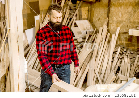 Craftsman in a woodworking shop selects timber while surrounded by raw materials, showcasing skill and creativity in a rustic setting Craftsman in a woodworking shop selects timber while surrounded by raw materials, showcasing skill and creativity in a rustic setting 130582191