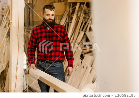 Woodworker carefully handles lumber in a workshop surrounded by tools and timber at daytime 130582193