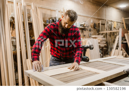 Skilled carpenter works on crafting a wooden door in a workshop during daylight hours 130582196