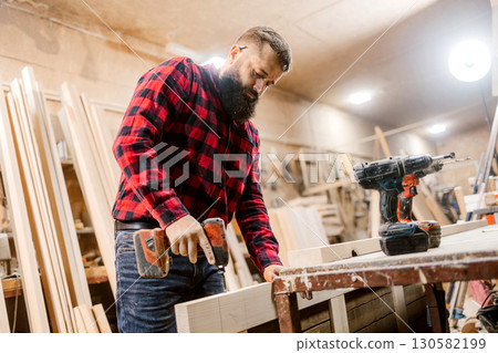 Skilled carpenter working in a workshop using a power drill to build wooden furniture Skilled carpenter working in a workshop using a power drill to build wooden furniture 130582199