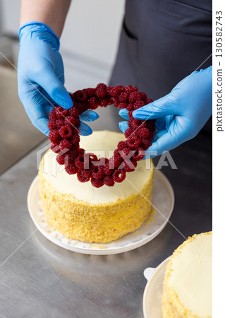 Chef decorating cake with fresh raspberry wreath on top in professional bakery kitchen Chef decorating cake with fresh raspberry wreath on top in professional bakery kitchen 130582743
