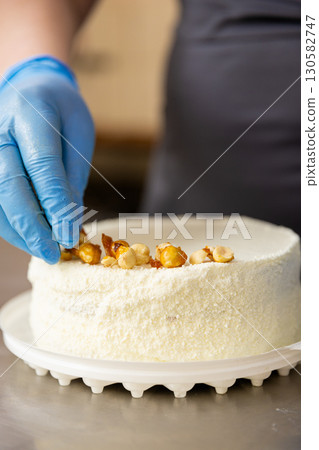 Pastry chef decorating white hazelnut cake by hand in food production kitchen Pastry chef decorating white hazelnut cake by hand in food production kitchen 130582747