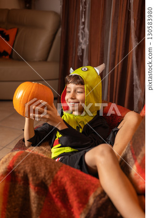 Happy child wearing a playful monster costume, holding a bright pumpkin while sitting comfortably on a sofa in a festively decorated living room, celebrating halloween at home Happy child wearing a playful monster costume, holding a bright pumpkin while sitting comfortably on a sofa in a festively decorated living room, celebrating halloween at home 130583980