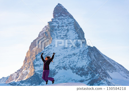 Happy woman in ski suit jumping outdoors on background of Matterhorn rock Happy woman in ski suit jumping outdoors on background of Matterhorn rock 130584218