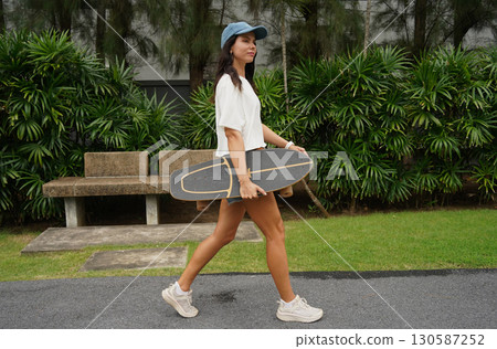 A woman is walking at the city park while carrying a skateboard in her hands 130587252