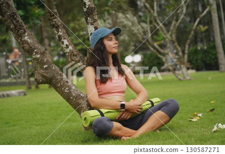 A young woman happily prepares to do yoga on a mat in a beautiful park. 130587271