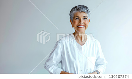 Smiling elderly woman in white shirt standing against light background Smiling elderly woman in white shirt standing against light background 130587274