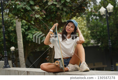 A young woman sits comfortably on a low wall at the park holding a skateboard in her hands 130587278