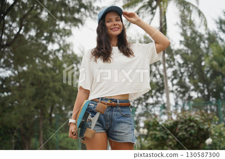 A woman is walking at the city park while carrying a skateboard in her hands A woman is walking at the city park while carrying a skateboard in her hands 130587300