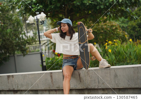 A young woman sits comfortably on a low wall at the park holding a skateboard in her hands 130587465