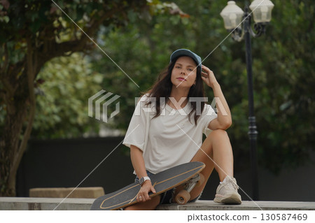 A young woman sits comfortably on a low wall at the park holding a skateboard in her hands A young woman sits comfortably on a low wall at the park holding a skateboard in her hands 130587469