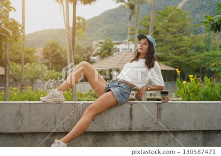 A young woman sits comfortably on a low wall at the park holding a skateboard in her hands 130587471
