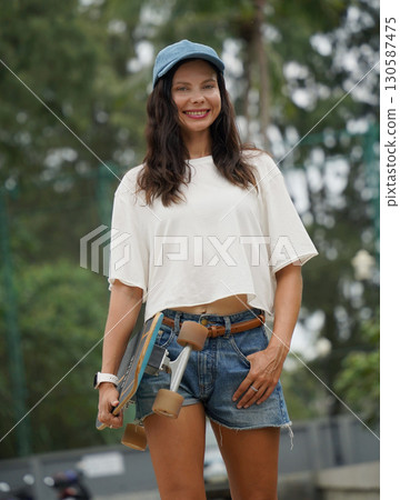 A woman is walking at the city park while carrying a skateboard in her hands A woman is walking at the city park while carrying a skateboard in her hands 130587475