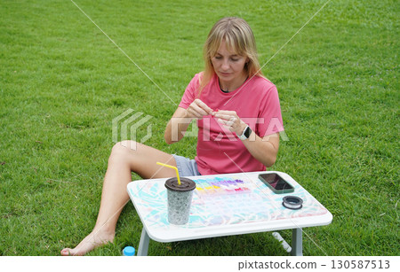 Young woman beading colorful bracelets outdoors in a park 130587513
