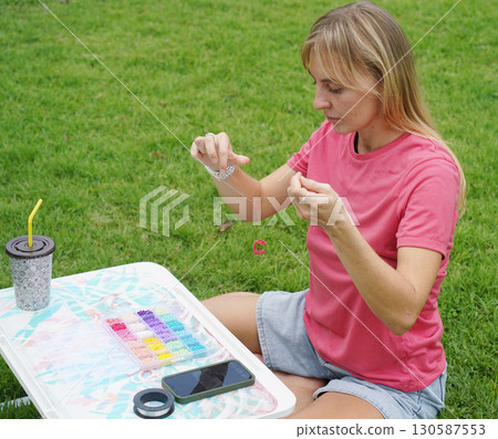 Young woman beading colorful bracelets outdoors in a park 130587553