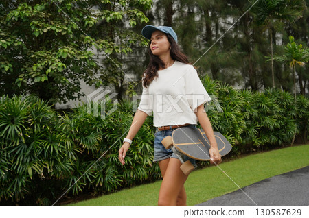 A woman is walking at the city park while carrying a skateboard in her hands A woman is walking at the city park while carrying a skateboard in her hands 130587629