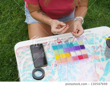 Young woman beading colorful bracelets outdoors in a park 130587699