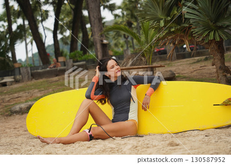 Woman in stylish wetsuit posing confidently with surfboard on beach Woman in stylish wetsuit posing confidently with surfboard on beach 130587952