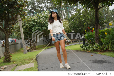 A woman is walking at the city park while carrying a skateboard in her hands A woman is walking at the city park while carrying a skateboard in her hands 130588107