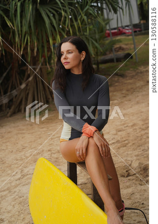 Woman in stylish wetsuit posing confidently with surfboard on beach Woman in stylish wetsuit posing confidently with surfboard on beach 130588116