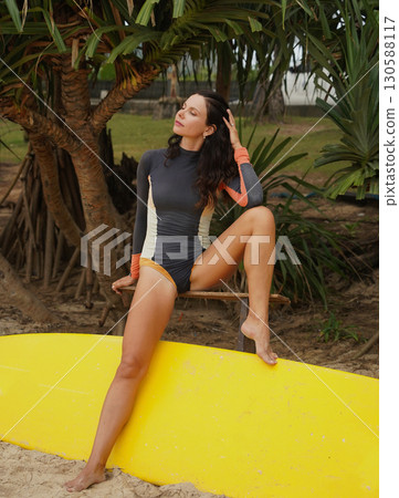 Woman in stylish wetsuit posing confidently with surfboard on beach 130588117