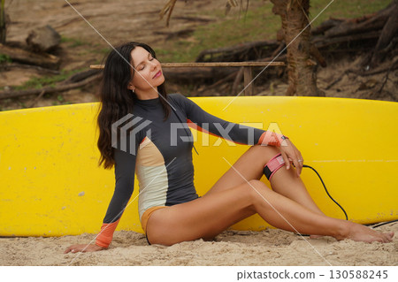 Woman in stylish wetsuit posing confidently with surfboard on beach 130588245