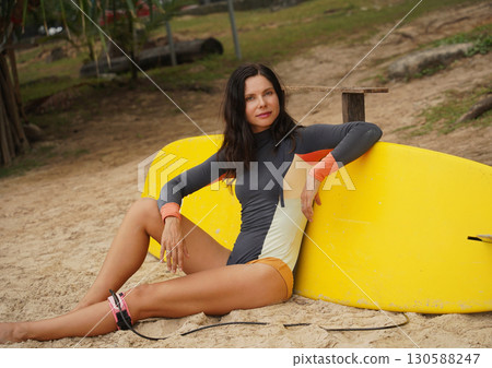 Woman in stylish wetsuit posing confidently with surfboard on beach 130588247