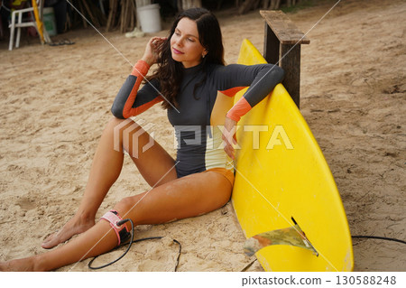 Woman in stylish wetsuit posing confidently with surfboard on beach Woman in stylish wetsuit posing confidently with surfboard on beach 130588248