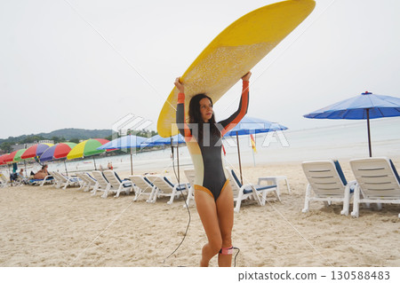 Young woman wearing tight wetsuit is going to surfboard at beautiful beach 130588483