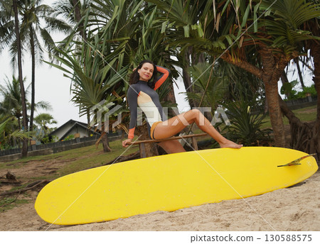 Woman in stylish wetsuit posing confidently with surfboard on beach Woman in stylish wetsuit posing confidently with surfboard on beach 130588575