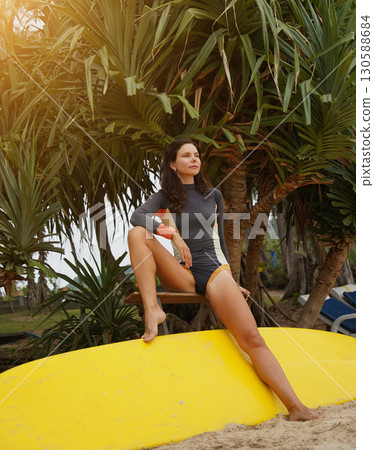 Woman in stylish wetsuit posing confidently with surfboard on beach Woman in stylish wetsuit posing confidently with surfboard on beach 130588684