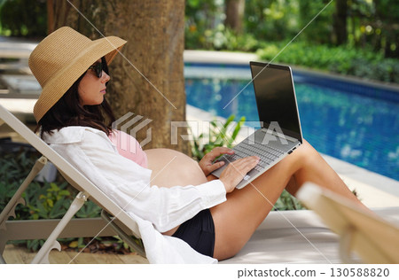 A pregnant woman is comfortably seated on a lounge chair using her laptop A pregnant woman is comfortably seated on a lounge chair using her laptop 130588820