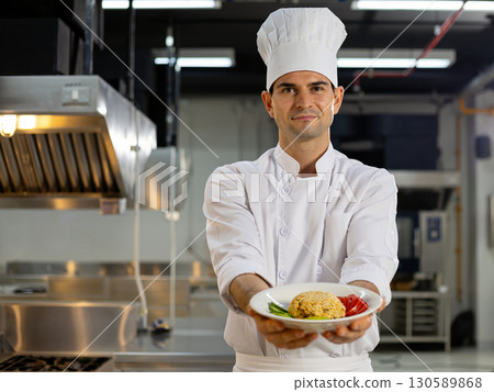 Smiling professional chef wearing white uniform and hat holding a plate of fried rice with vegetables inside a commercial kitchen Smiling professional chef wearing white uniform and hat holding a plate of fried rice with vegetables inside a commercial kitchen 130589868